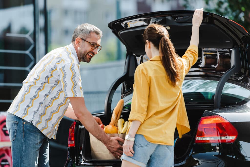 Selective focus of woman opening car trunk near smiling man holding shopping bag with food on urban street