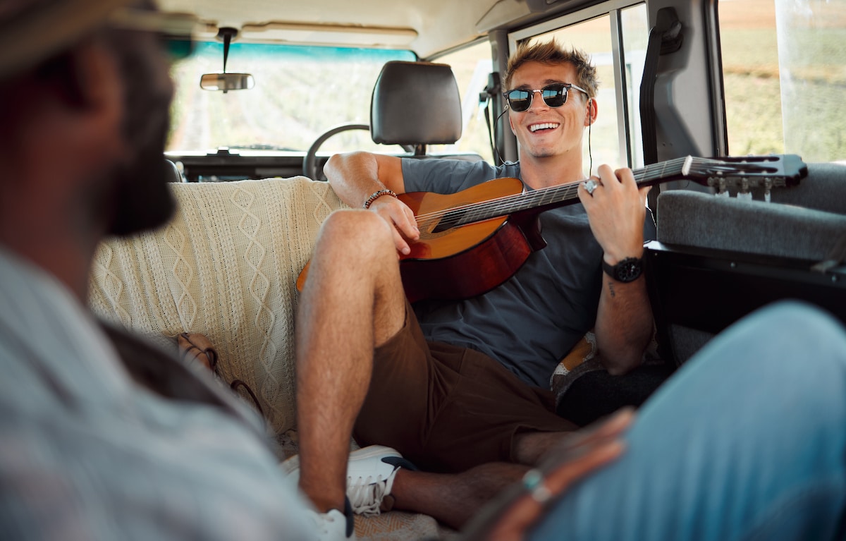 A smiling young man playing the guitar in a van, traveling with his friend.