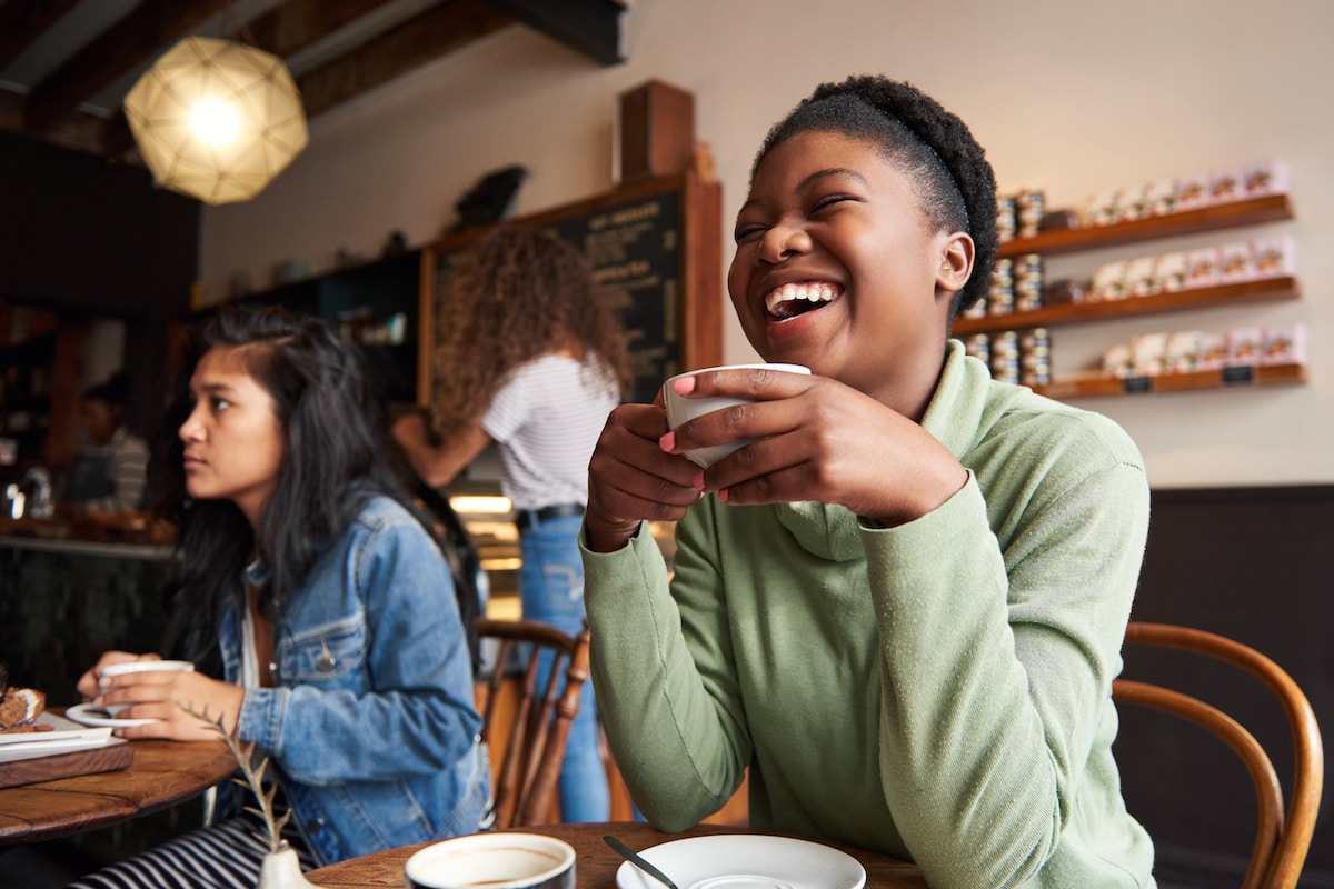 Young African American woman laughing while sitting in a cafe drinking coffee and hanging out with friends