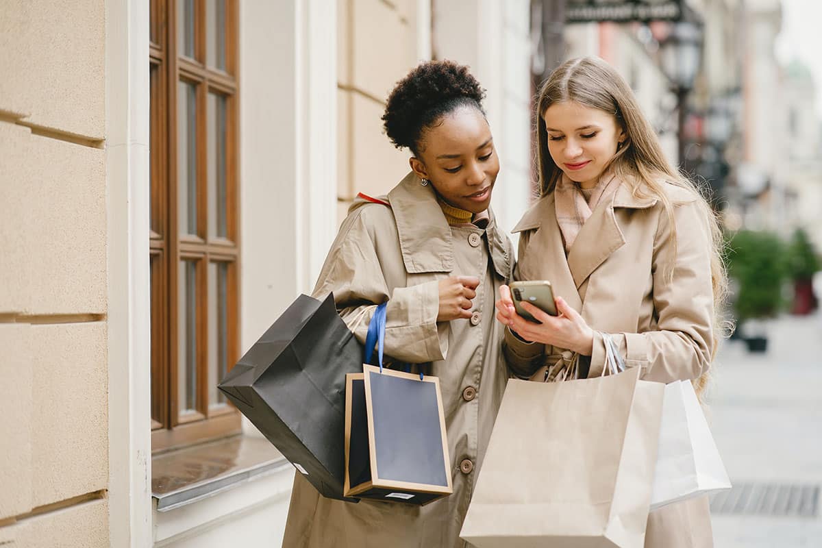two women outside looking at one of the women's phones while holding shopping bags